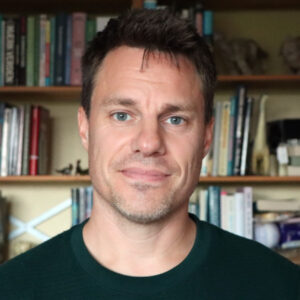 Foto af Anders Stjernholm foran en bogreol. Varmt smilende mand i 40'erne med kort mørkt hår og klare blågrønne øjne