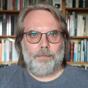 Foto af Claus Gregers Jørgensen foran en bogreol. Mand i 40'erne med sky smil, kraftig gråt skæg og skulderlangt hår.