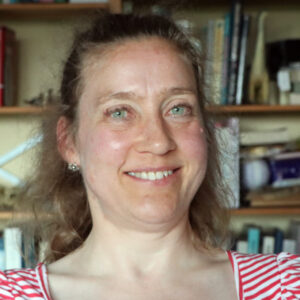 Foto af Nana Prentø foran en bogreol. Storsmilende kvinde i 50'erne med klare øjen og langt krøllet hår.