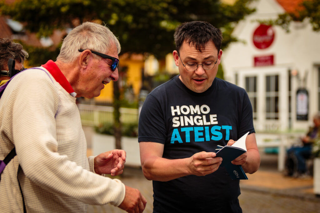 Medlem af Ateistisk Selskab Rasmus Malver viser den ateistiske bibel frem til begejstret ældre herre. Spoler alert... Den er tom. Du bestemmer selv, hvad du vil tro og tænke. På Rasmus t-shirt står homo, single, ateist.